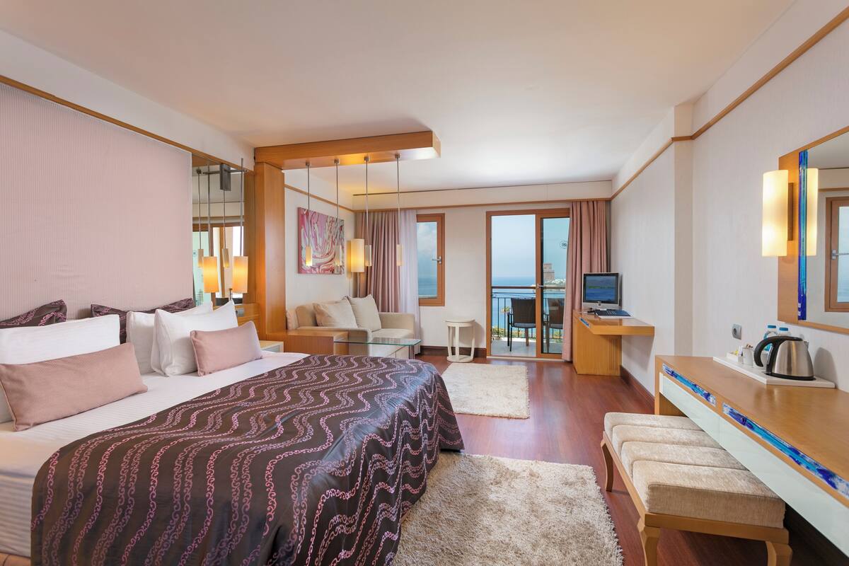 A hotel room with a large bed, a desk with a computer, a TV, and a balcony with a view.