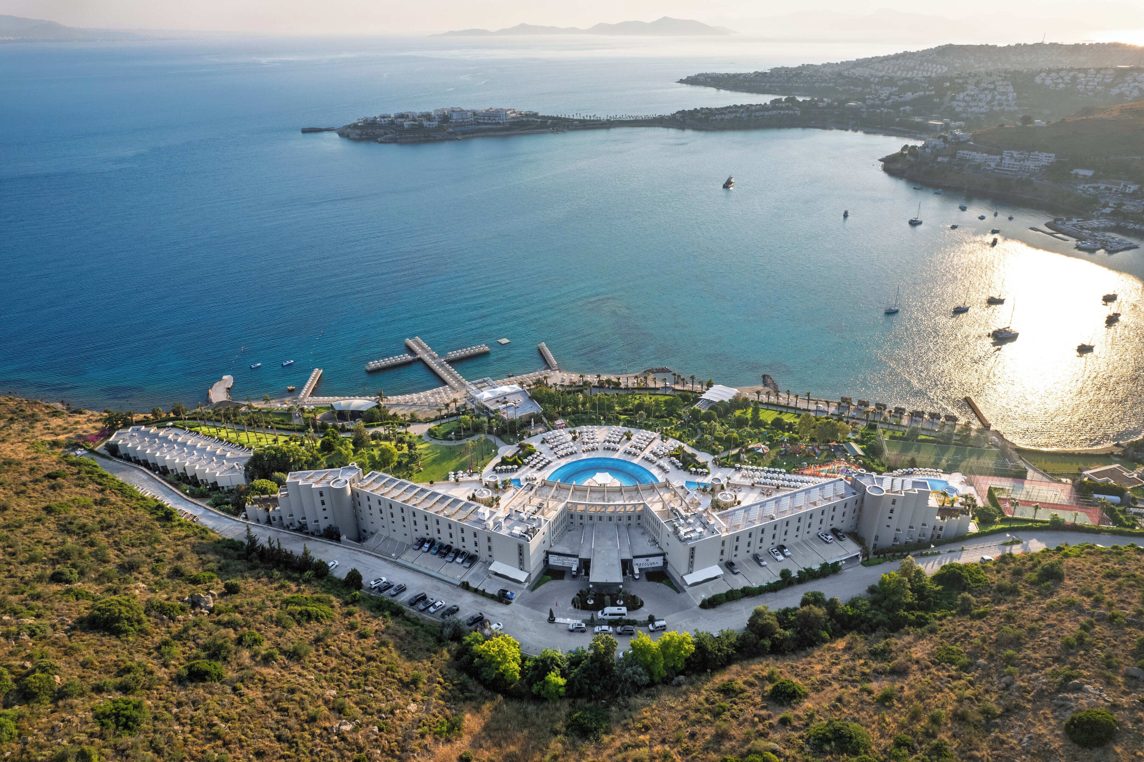 Um hotel grande com piscina central, várias áreas de estacionamento e vista para o mar.