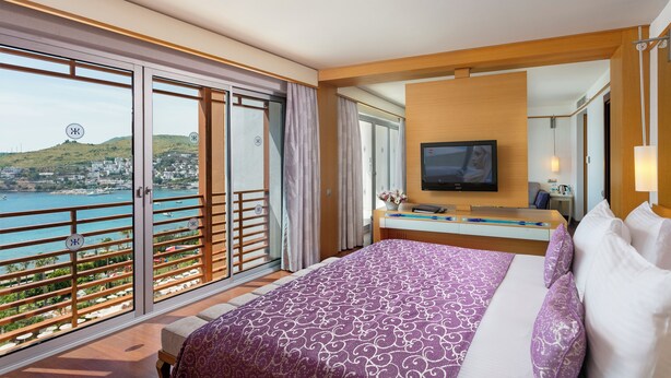 A hotel room with a bed, a TV, a desk, and a balcony with a view of the sea. - Kefaluka Resort - All Inclusive (Bodrum)