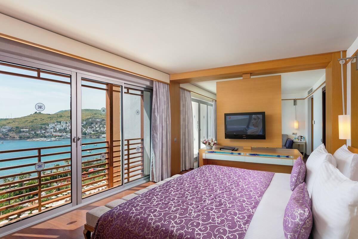 A hotel room with a bed, a TV, a desk, and a balcony with a view of the sea.