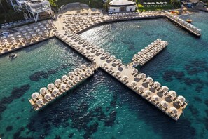 An aerial view of a resort with a central water body, connected by walkways with umbrellas, and surrounding beach areas with lounge chairs. - Kefaluka Resort - All Inclusive (Bodrum)