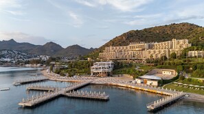 Un hotel frente al mar con un puerto deportivo, varias edificaciones y montañas al fondo.