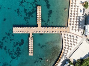 Vista aèria d'un moll amb files de gandules i ombrel·les, una zona de piscina amb ombrel·les i un mar blau i translúcid.