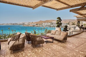 Una terraza con mobiliario de mimbre, un sofá y una mesa de centro. Se observa un pueblo costero con edificios y un mar azul cristalino.