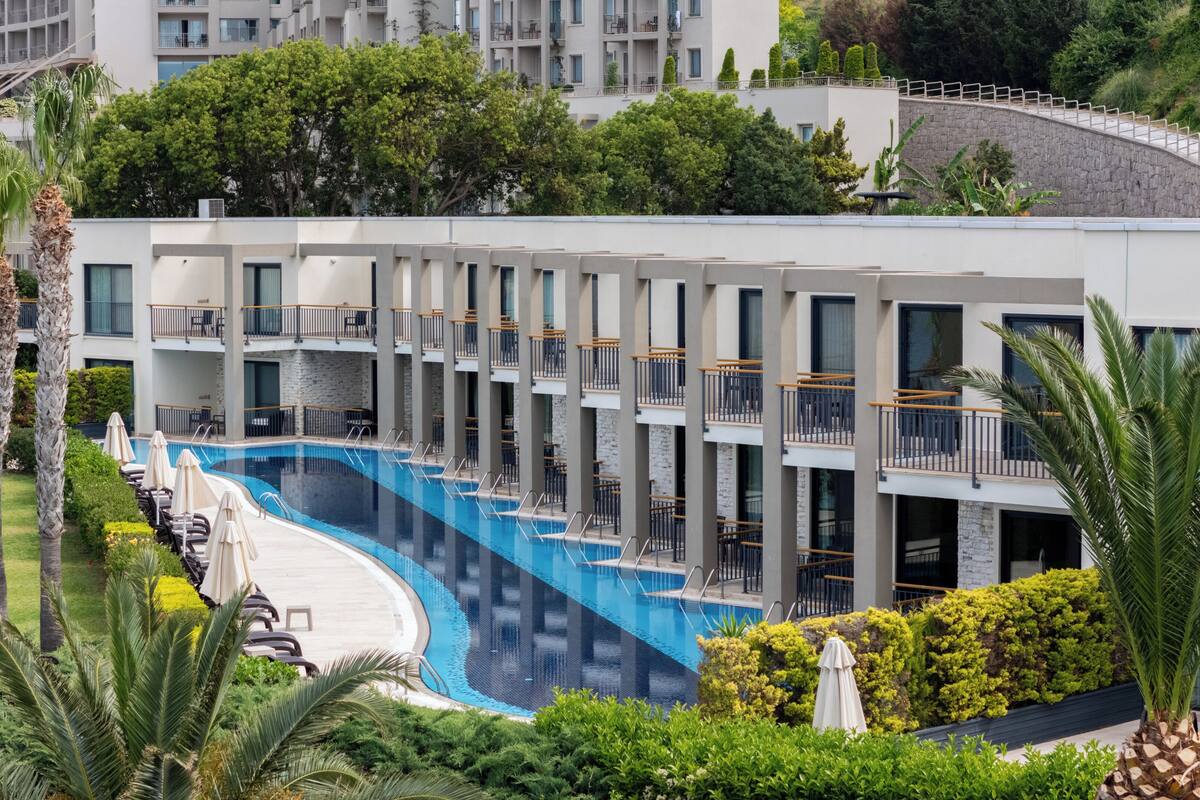 A modern hotel with a swimming pool, multiple balconies, and lush greenery.