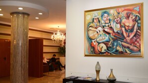 A lobby with a large marble column, a chandelier, a colorful painting, and a reception desk with a person sitting. - Iacone (Chieti)