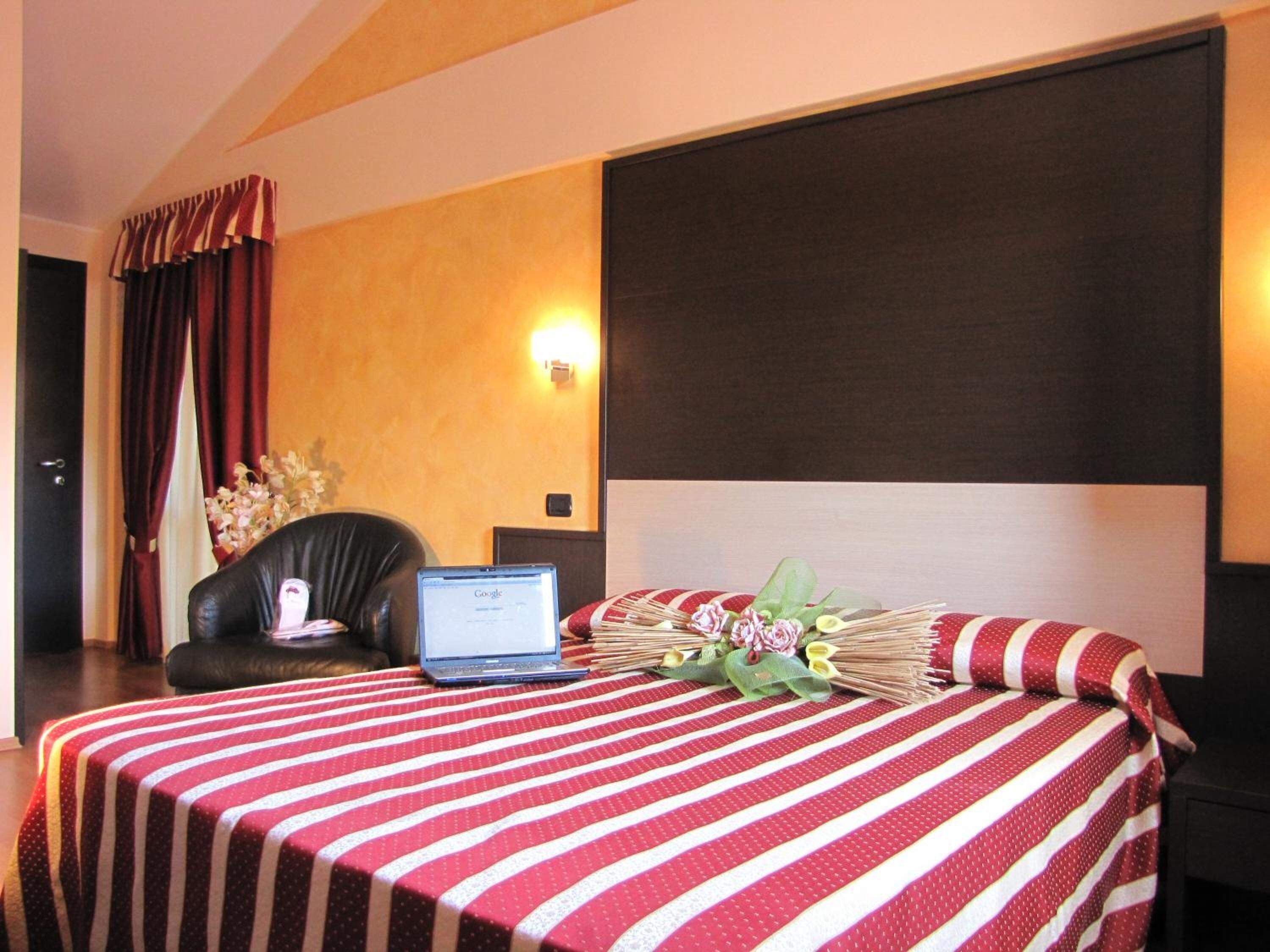 Habitación de hotel con cama, una laptop sobre una mesita de noche, una silla y una lámpara fijada a la pared.