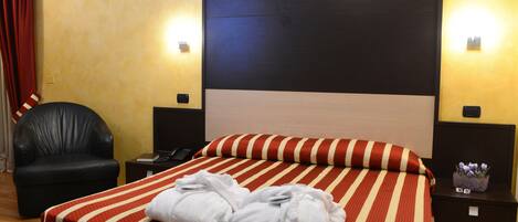 Habitación de hotel con una cama grande, un sillón de cuero negro, una mesita de noche con un teléfono y una pequeña mesa con un jarrón de flores.