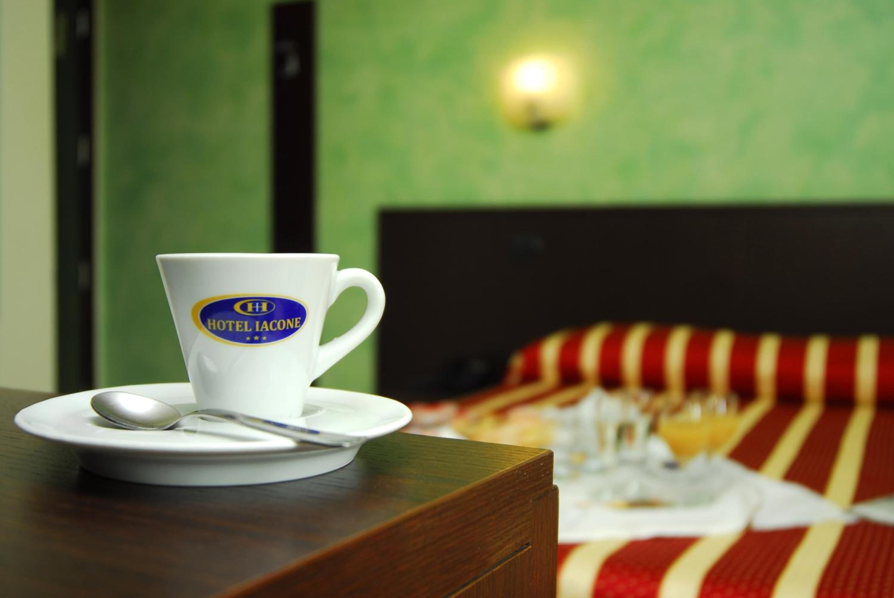 Eine weiße Tasse mit einem Logo auf einem Holztisch, daneben ein Löffel, im Hintergrund ein Bett mit einer gestreiften Decke.