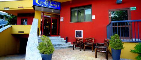 A hotel entrance with a red facade, a blue sign, and a set of stairs leading to the entrance. There are two potted plants and a bench with chairs outside.