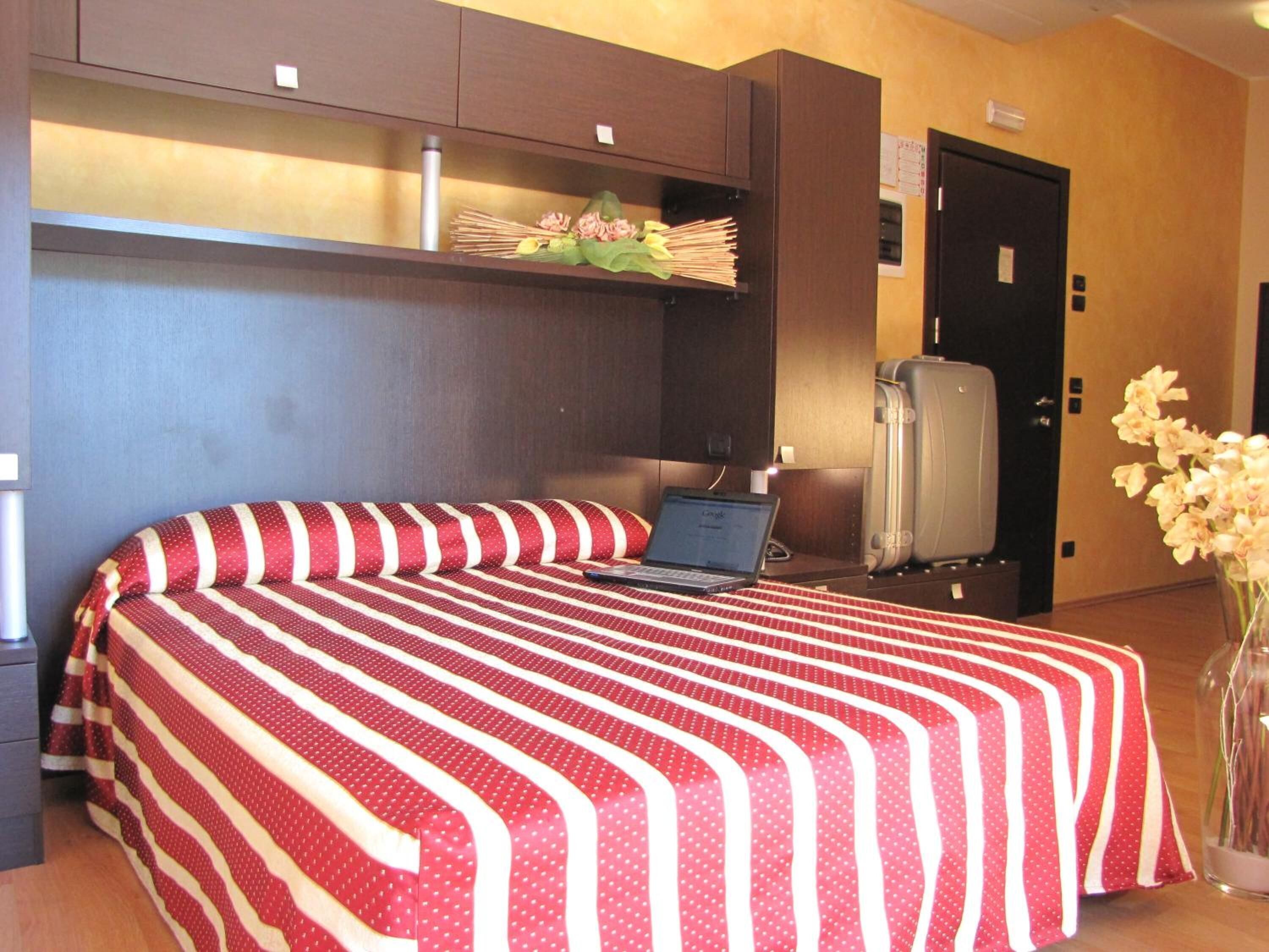 Une chambre d’hôtel avec un lit, un ordinateur portable posé sur le lit, un réfrigérateur et un vase rempli de fleurs.