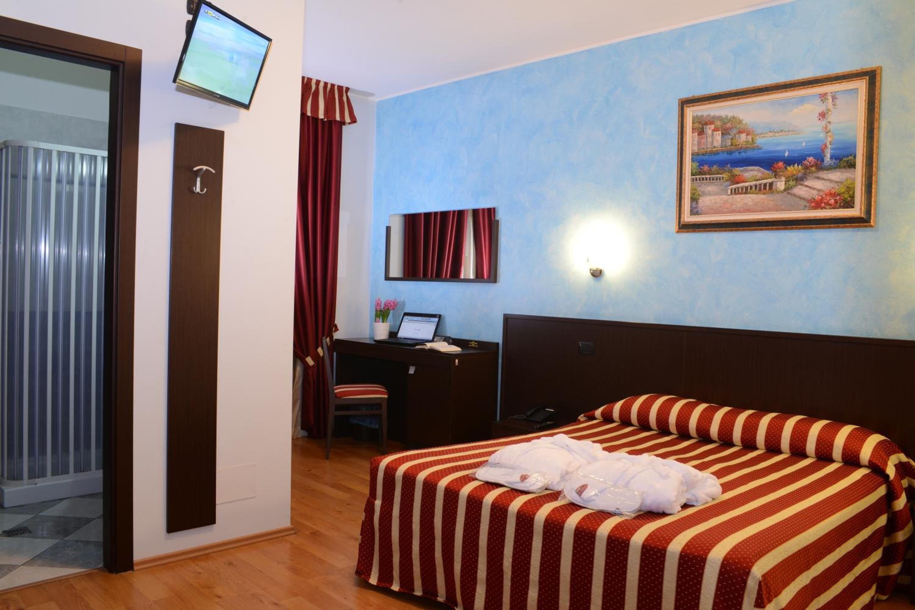 A hotel room with a bed, a desk, a computer, and a painting on the wall.