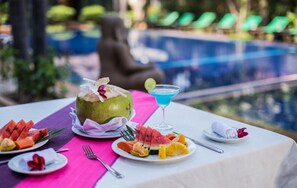 Una mesa con un postre de coco, rebanadas de sandía y un cóctel azul, con vista a la piscina.