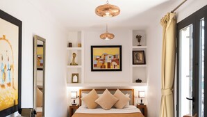 A hotel room with a bed, bedside tables, a desk with a chair, a mirror, artwork on the walls, and a view through a window with curtains.