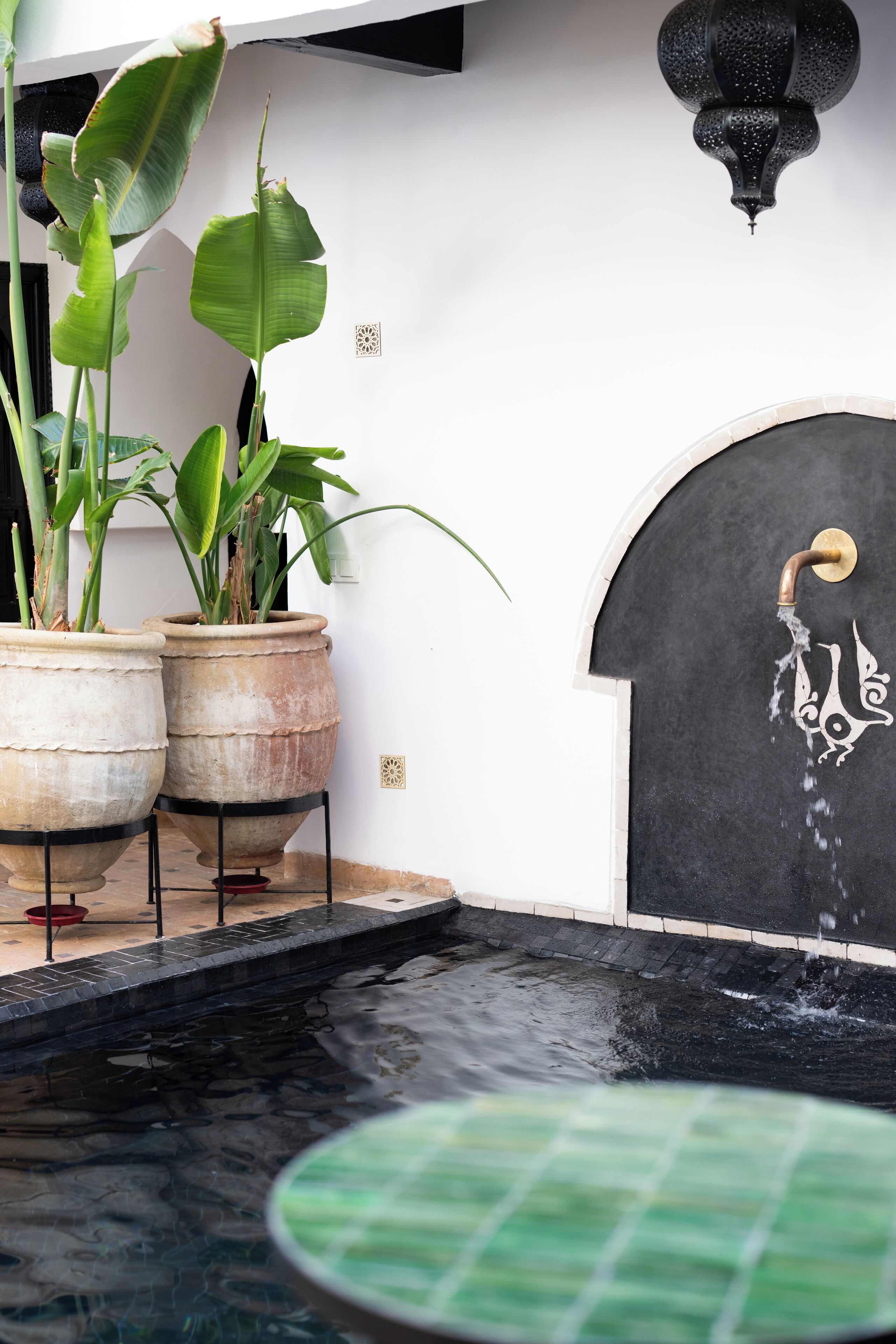 Zona de piscina con fuente, plantas en macetas y un muro decorativo.
