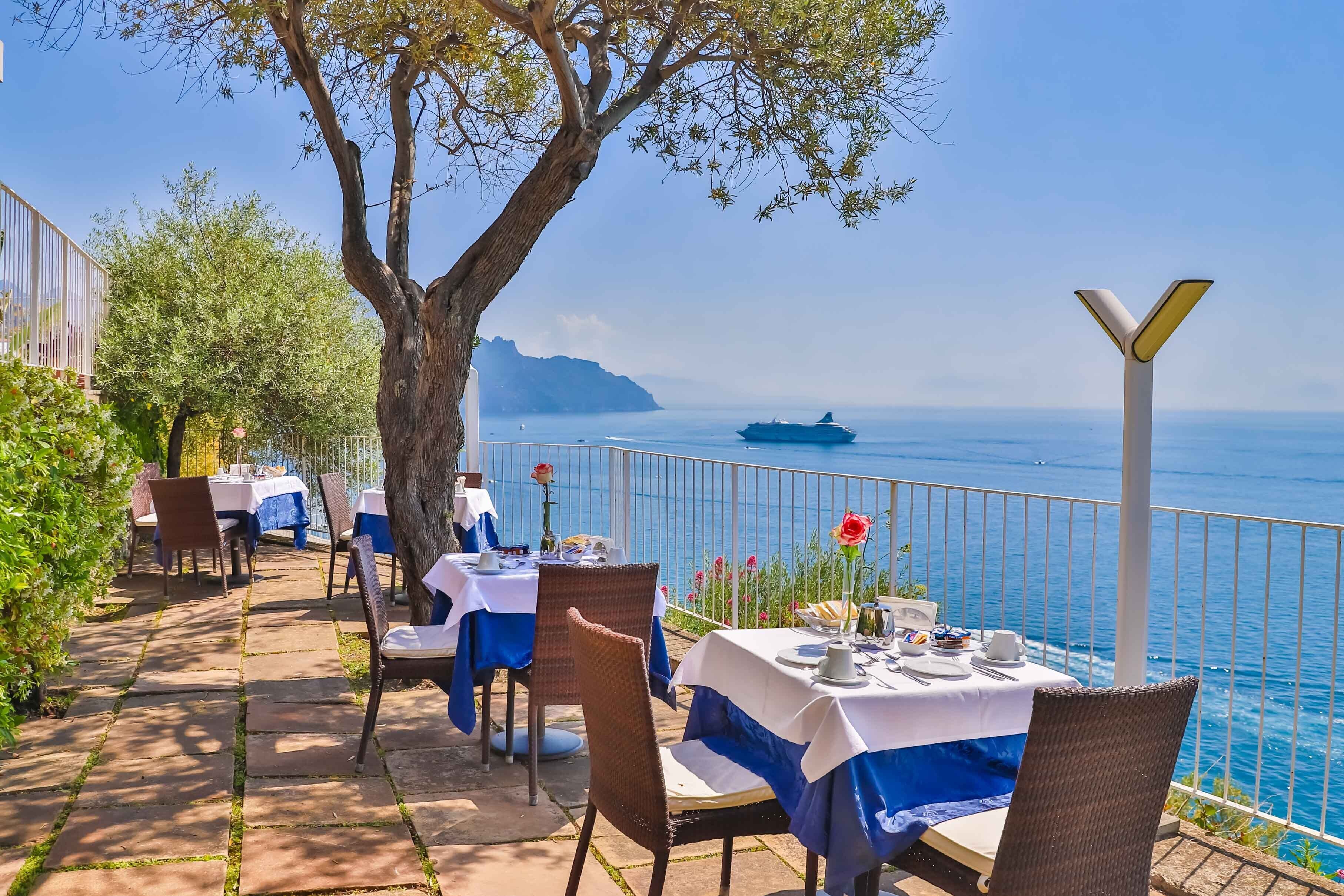 An outdoor dining area with tables set for two, overlooking a body of water with a ship in the distance.