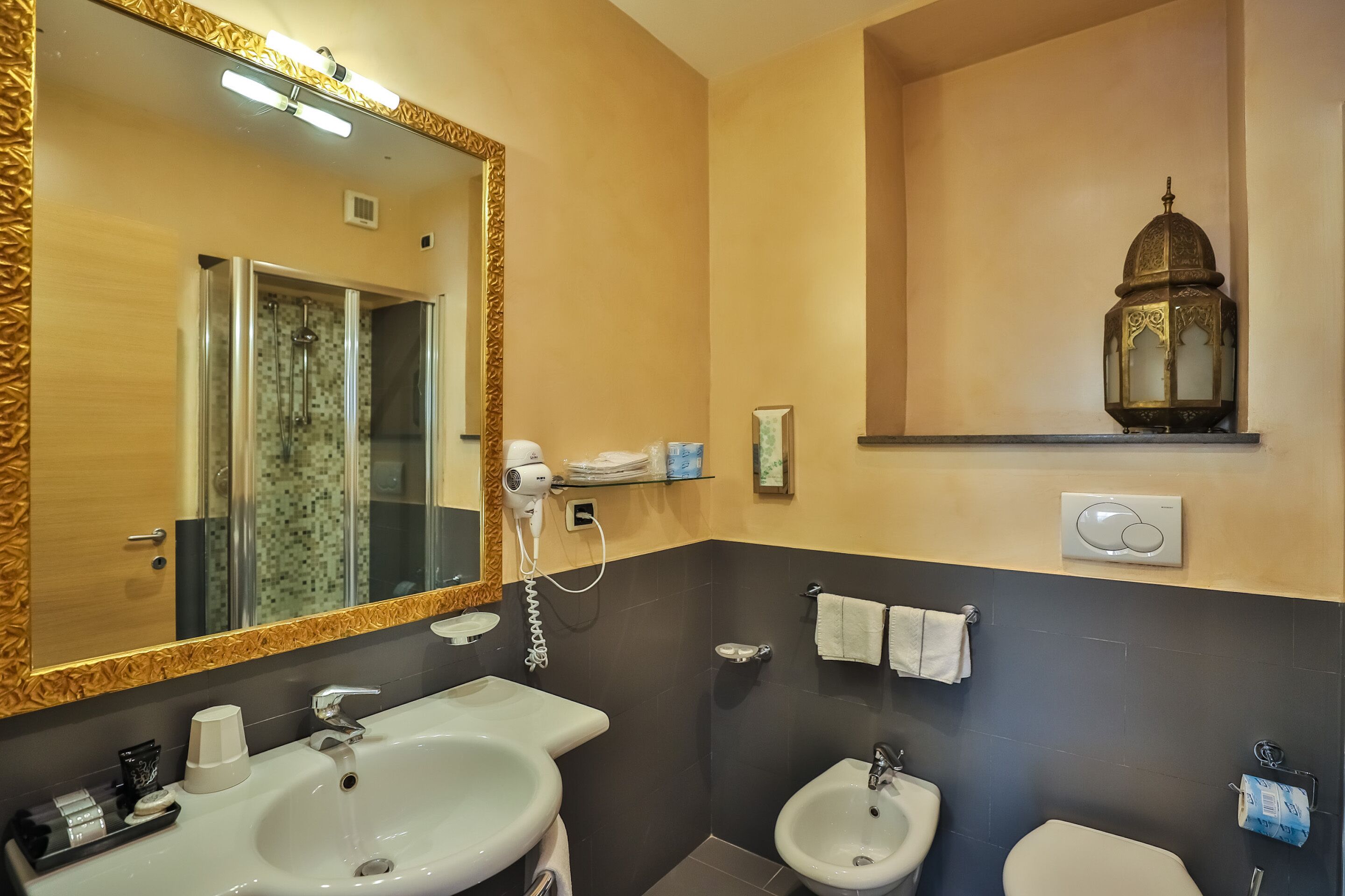 A bathroom with a large mirror, a sink, a bidet, and a toilet. There is a hairdryer on the wall and a decorative lantern above the toilet.