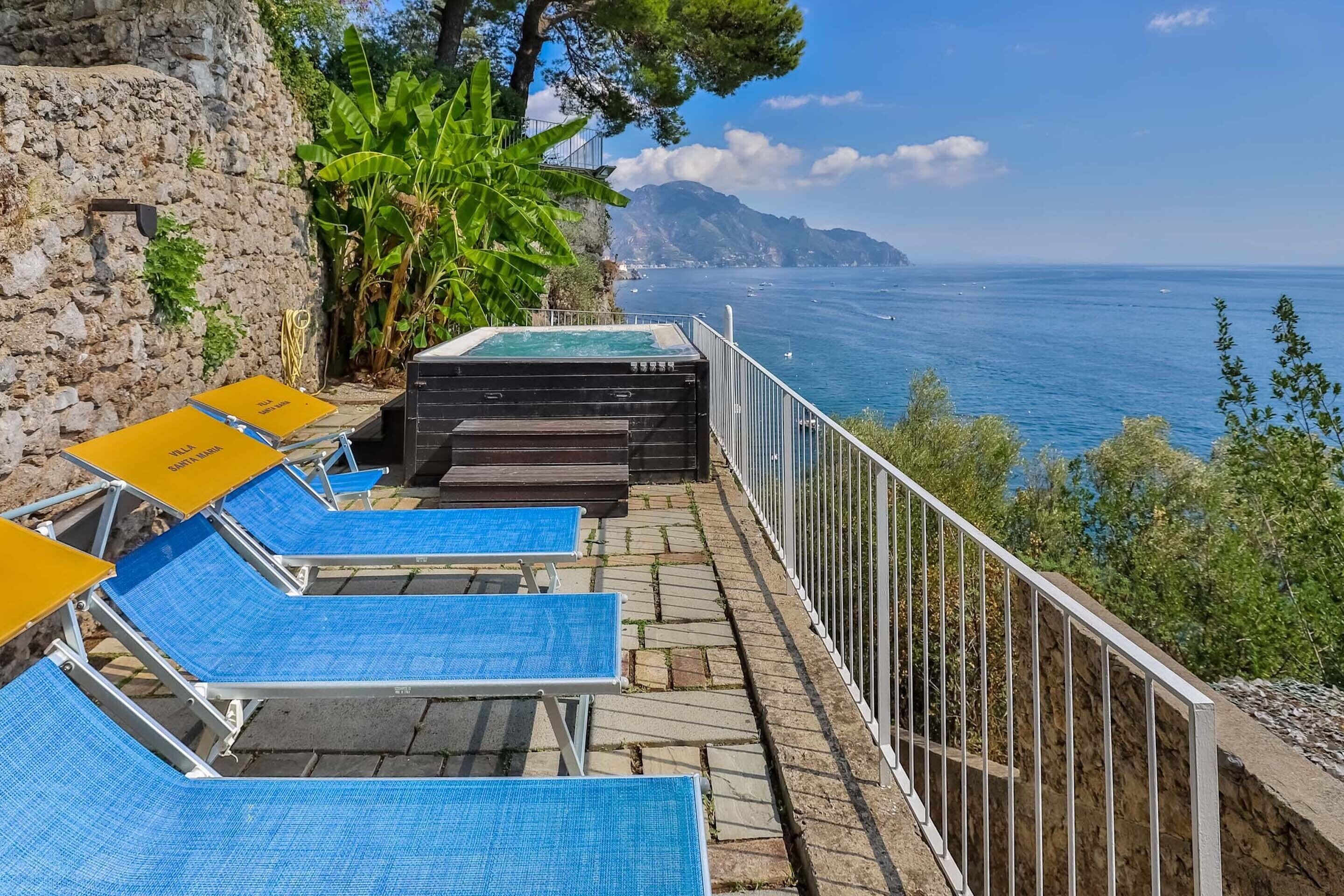 Una terraza de piedra con tumbonas azules y una bañera de hidromasaje con vistas al mar.