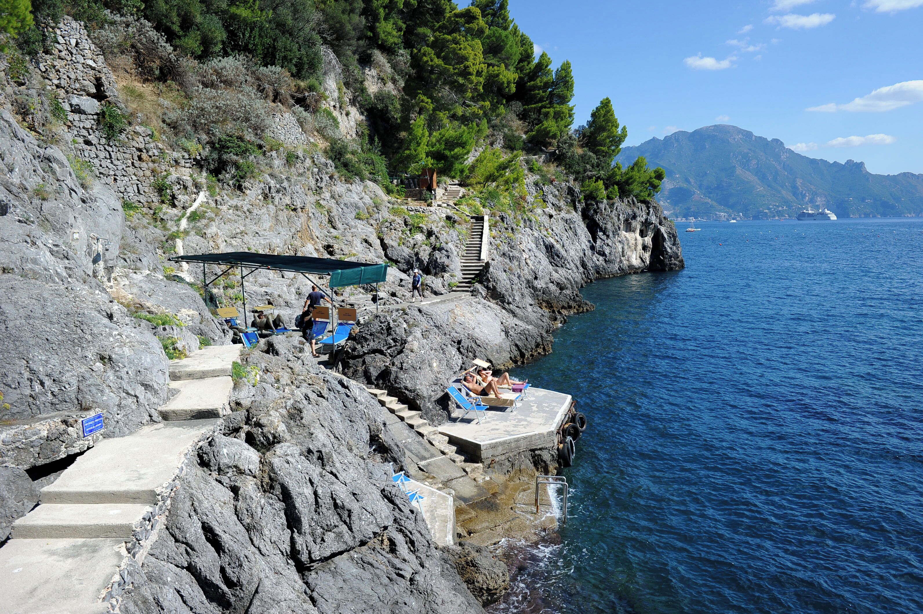 A rocky coastal area with steps leading down to the sea, a small dock with a boat, and a few people relaxing by the water.