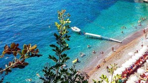 Escena costera con aguas azules cristalinas, barcos y una playa con gente.