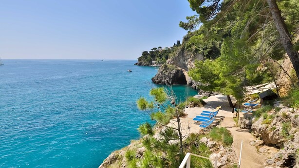 Una zona costera con mar azul cristalino, un sendero pedregoso y sillas de descanso azules.