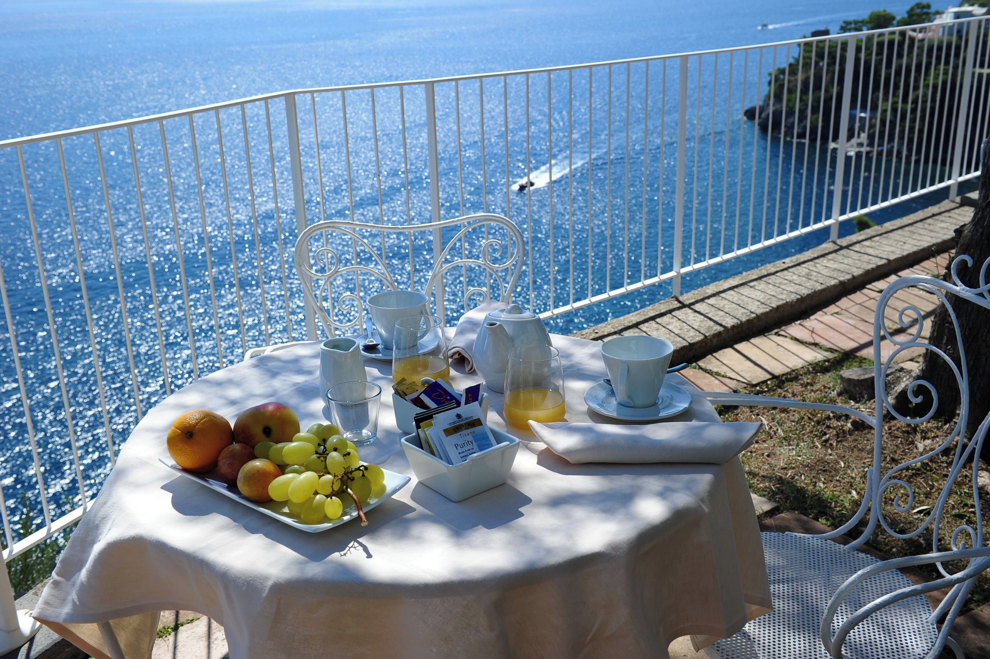 Een tafel gedekt met fruit, bekers en een menukaart op een balkon met uitzicht op een waterpartij.