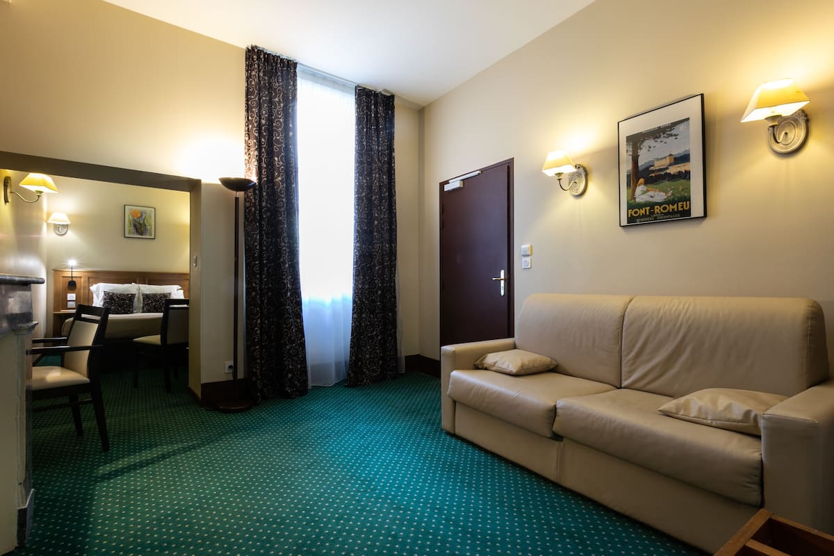 A hotel room with a green patterned carpet, a beige sofa, a dining area with a table and chairs, and two wall-mounted lamps.