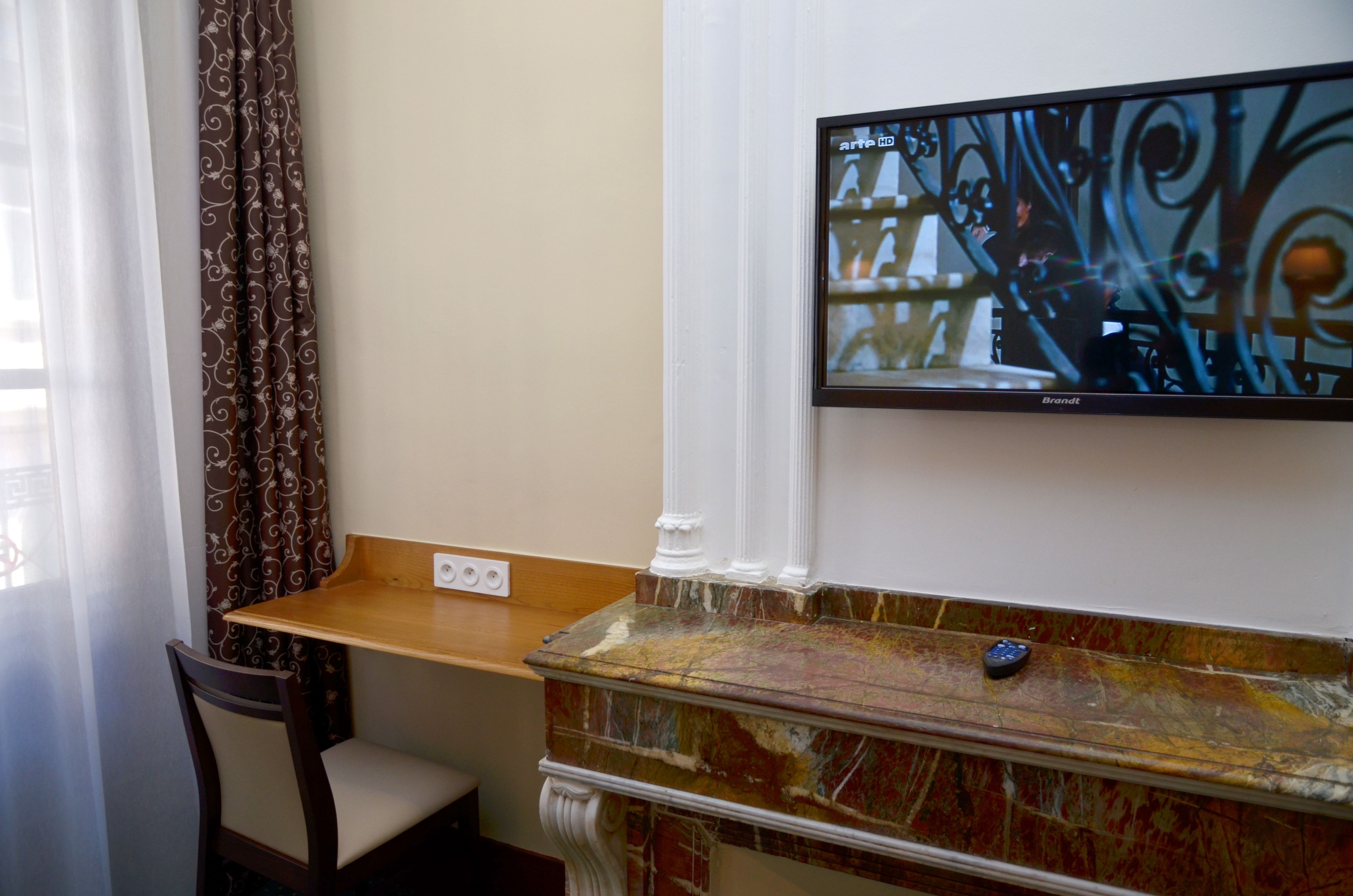A room with a wooden table, a chair, a television mounted on the wall, and a marble fireplace.