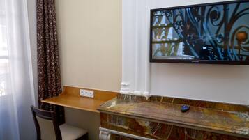 A room with a wooden table, a chair, a television mounted on the wall, and a marble fireplace.