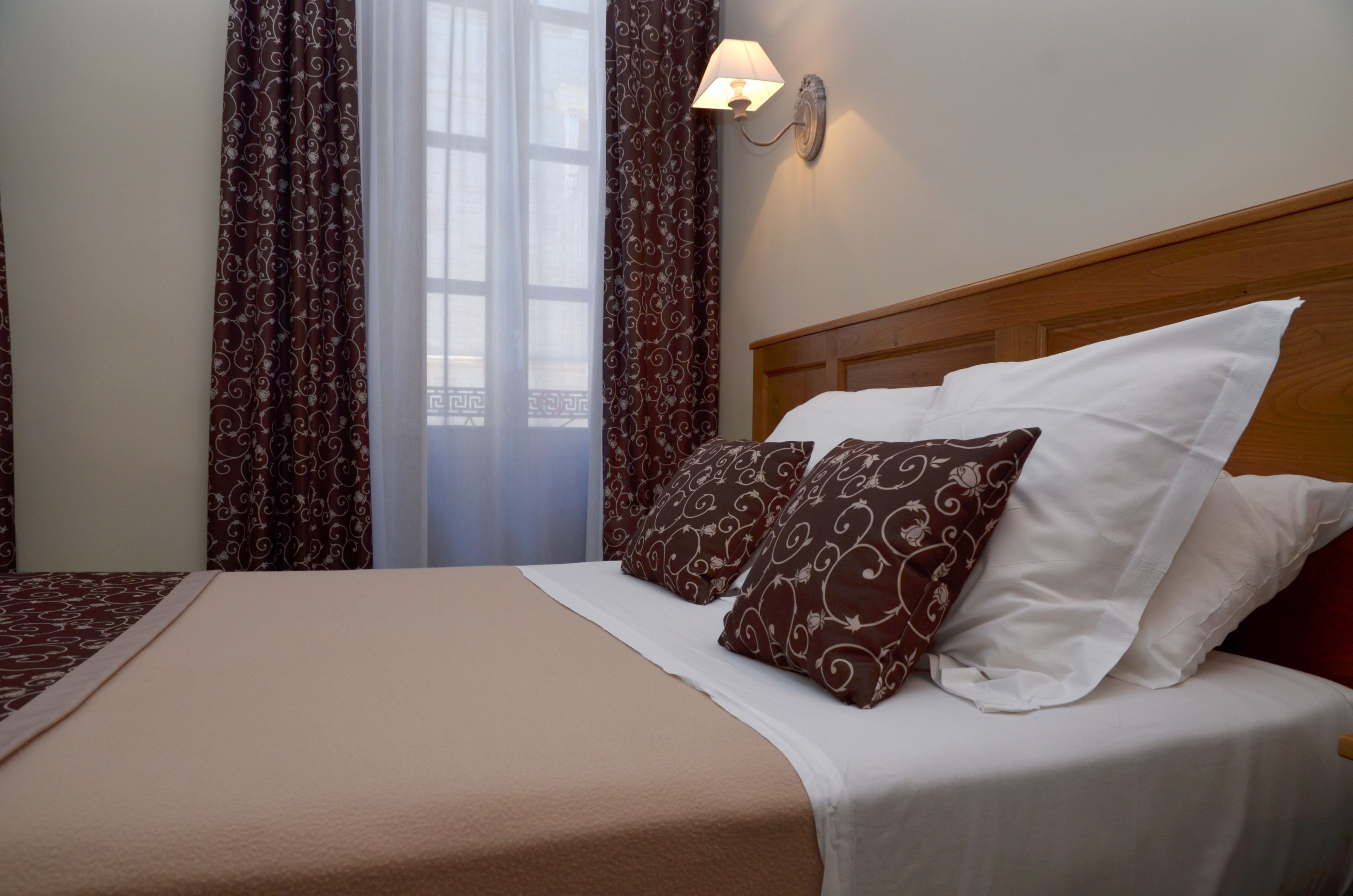 Une chambre d’hôtel avec un lit, des rideaux, une lampe fixée au mur et une fenêtre donnant sur un paysage.