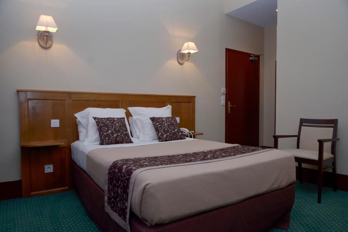 Quarto de hotel com cabeceira de madeira, cama com almofadas brancas e estampadas, mesa de cabeceira com abajur e uma cadeira.