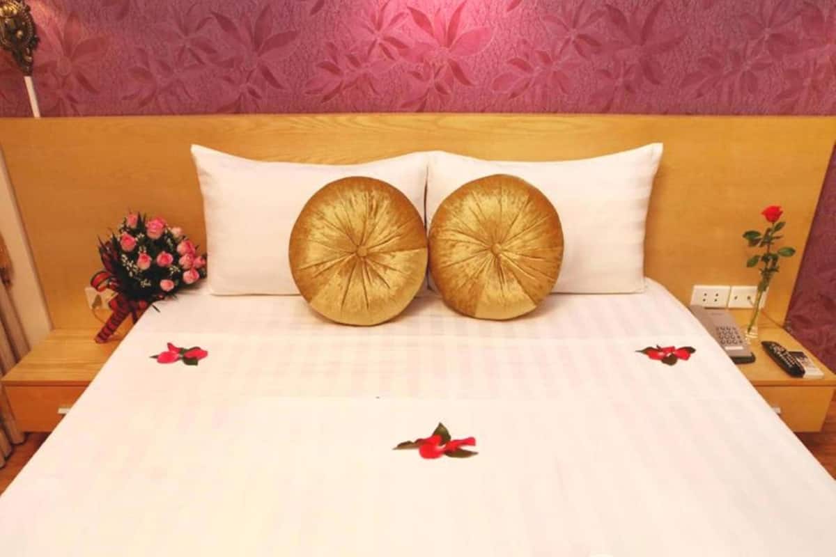 A hotel room with a bed, two golden pillows, a nightstand with a vase of flowers, and a remote control.