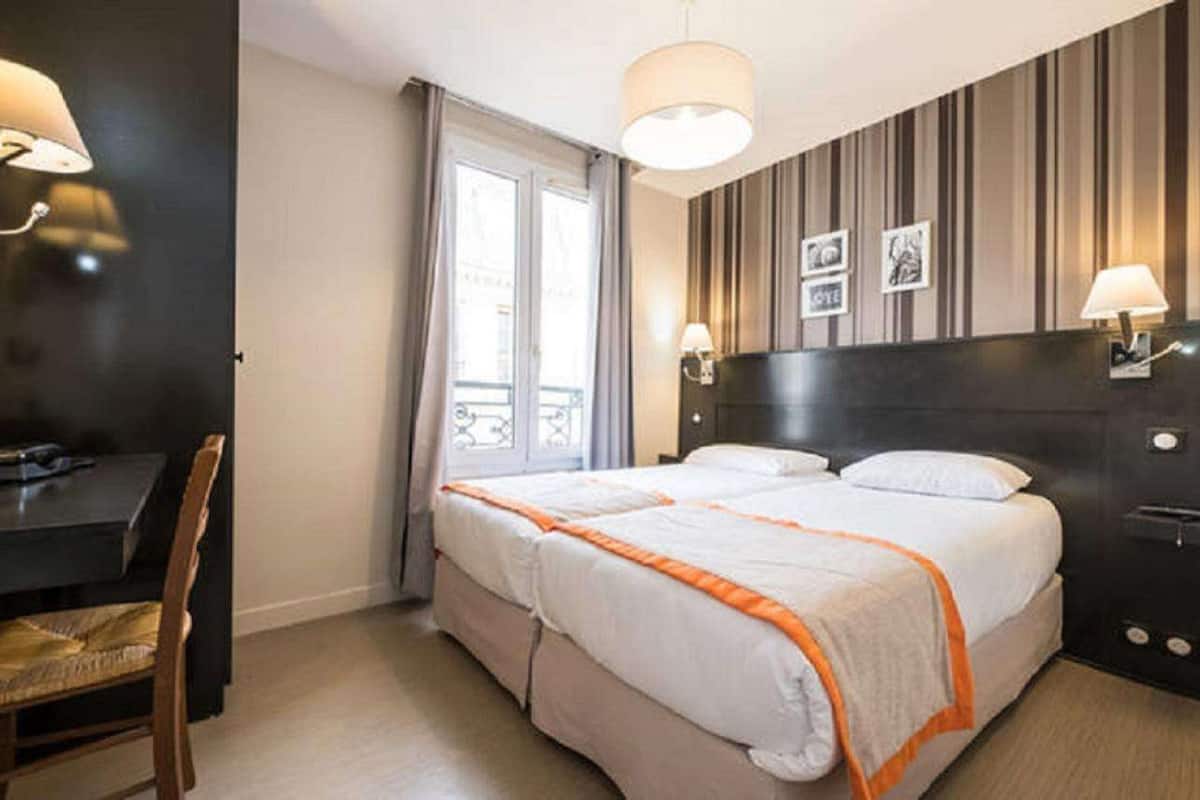 Une chambre d’hôtel avec un lit, un bureau et une chaise. Il y a deux lampes de chevet et une fenêtre avec des rideaux.