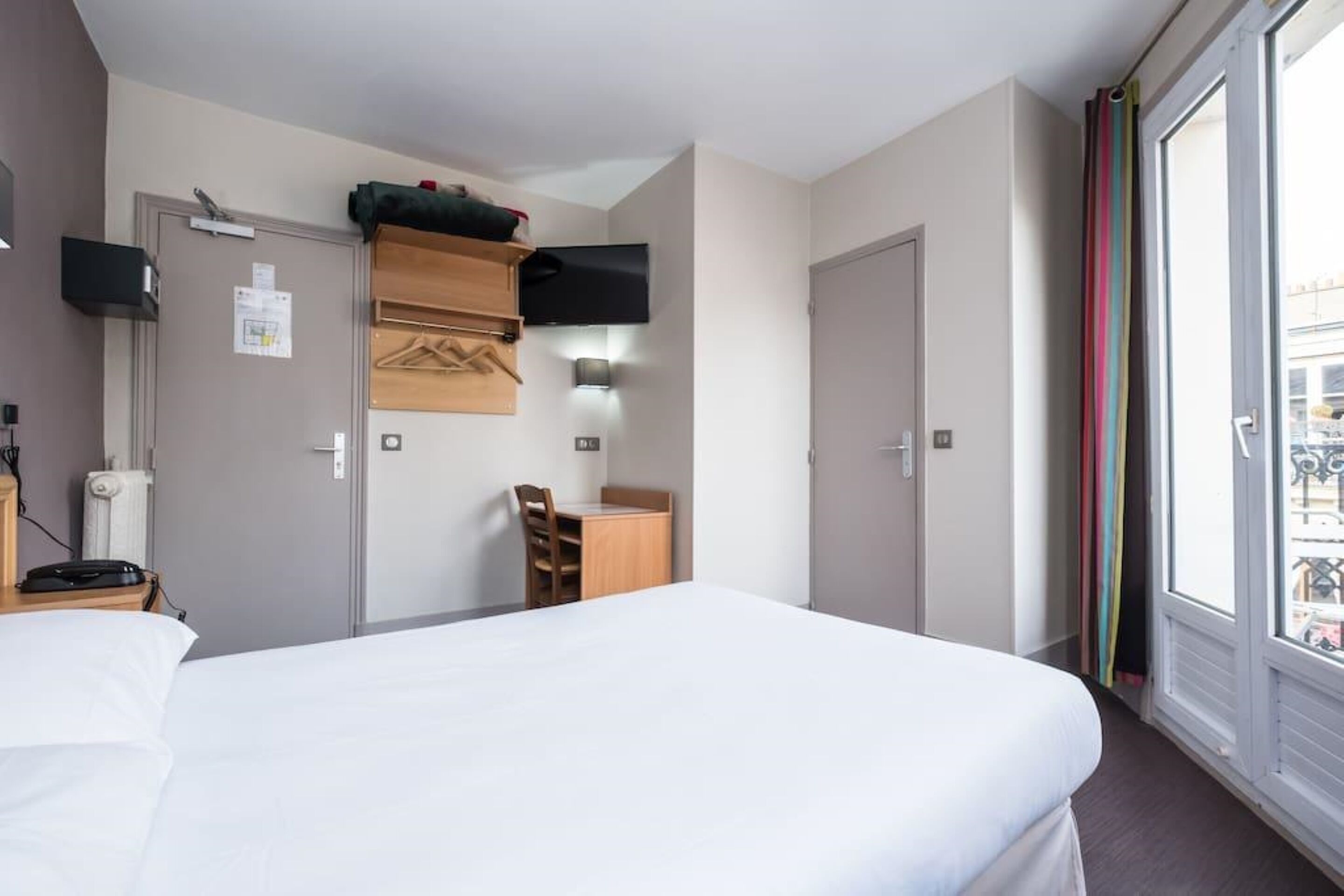 Une chambre d’hôtel dotée d’un grand lit, d’un bureau avec une chaise, d’un téléviseur à écran plat fixé au mur et d’une fenêtre agrémentée de rideaux colorés.