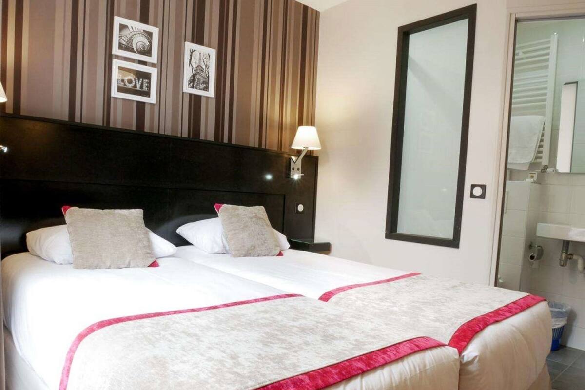 Une chambre d’hôtel avec deux lits, une tête de lit sombre, une lampe fixée au mur et un grand miroir.