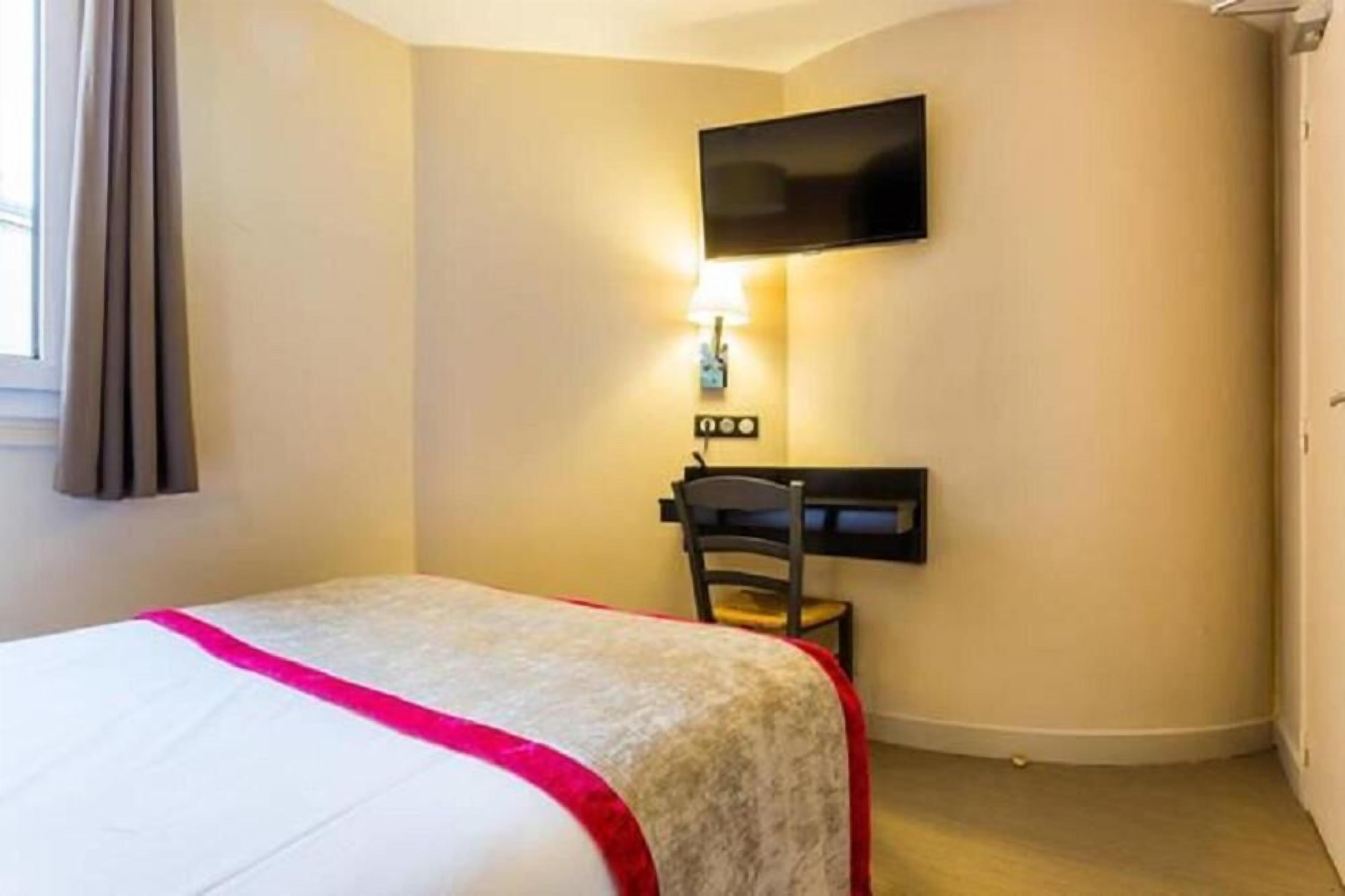 A hotel room with a bed, a desk, a chair, a TV mounted on the wall, and a window with curtains.