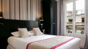 A hotel room with a large bed, striped wallpaper, a black headboard, a nightstand with a lamp, and a view of buildings through a window. - Hotel Bonsejour Montmartre (Paris)
