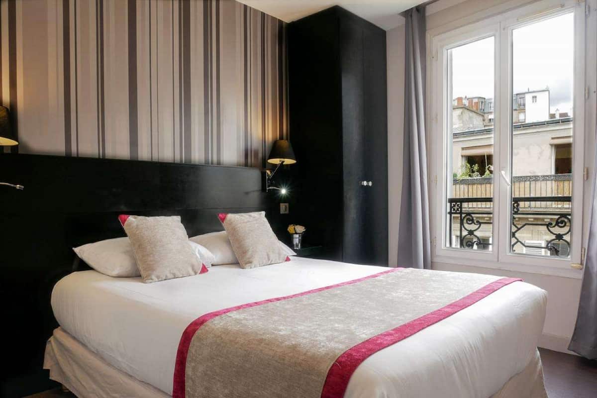 Une chambre d’hôtel avec un grand lit, un papier peint à rayures, une tête de lit noire, une table de chevet avec une lampe et une vue sur les bâtiments par la fenêtre.