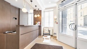 A hotel reception area with a wooden reception desk, a seating area with a chair and a small table, and a large glass door leading outside. - Hotel Bonsejour Montmartre (Paris)