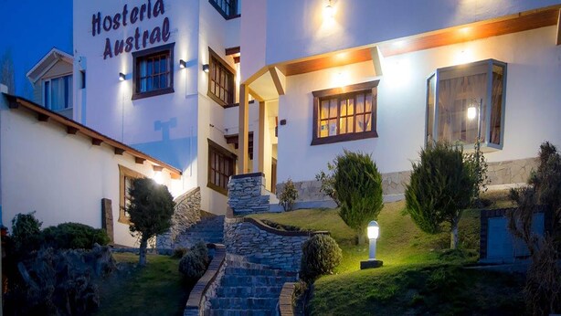 A two-story building with the sign "Hosteria Austral" at the entrance. There are stone steps leading up to the entrance, and a small garden area with a lamp post and trimmed bushes.