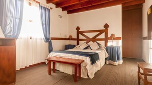 Un dormitorio con cama de madera, una mesita pequeña y una silla. Se observa una ventana con cortinas, un radiador y una puerta.