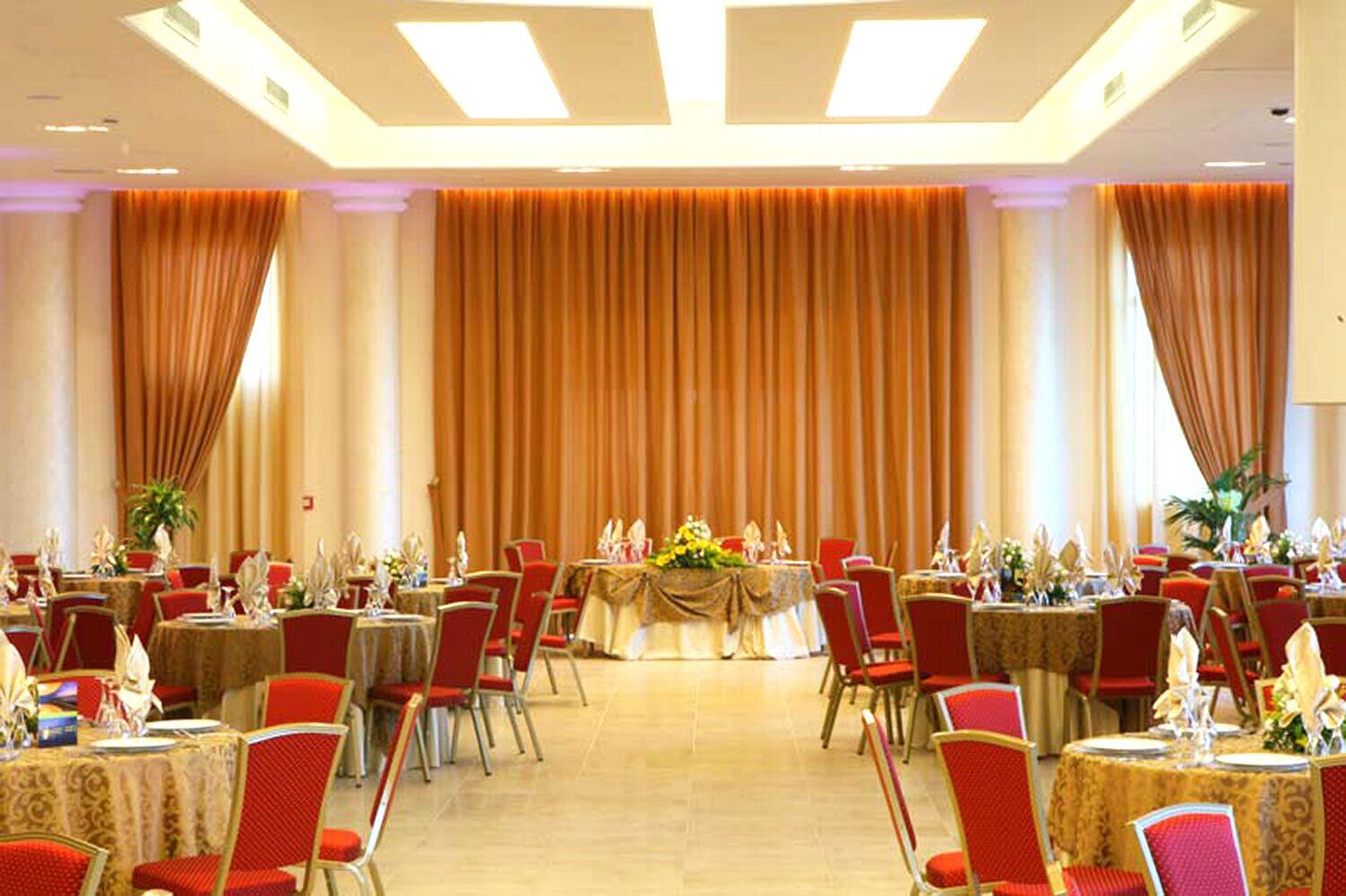 A banquet hall with round tables, red chairs, and a stage with a floral arrangement.