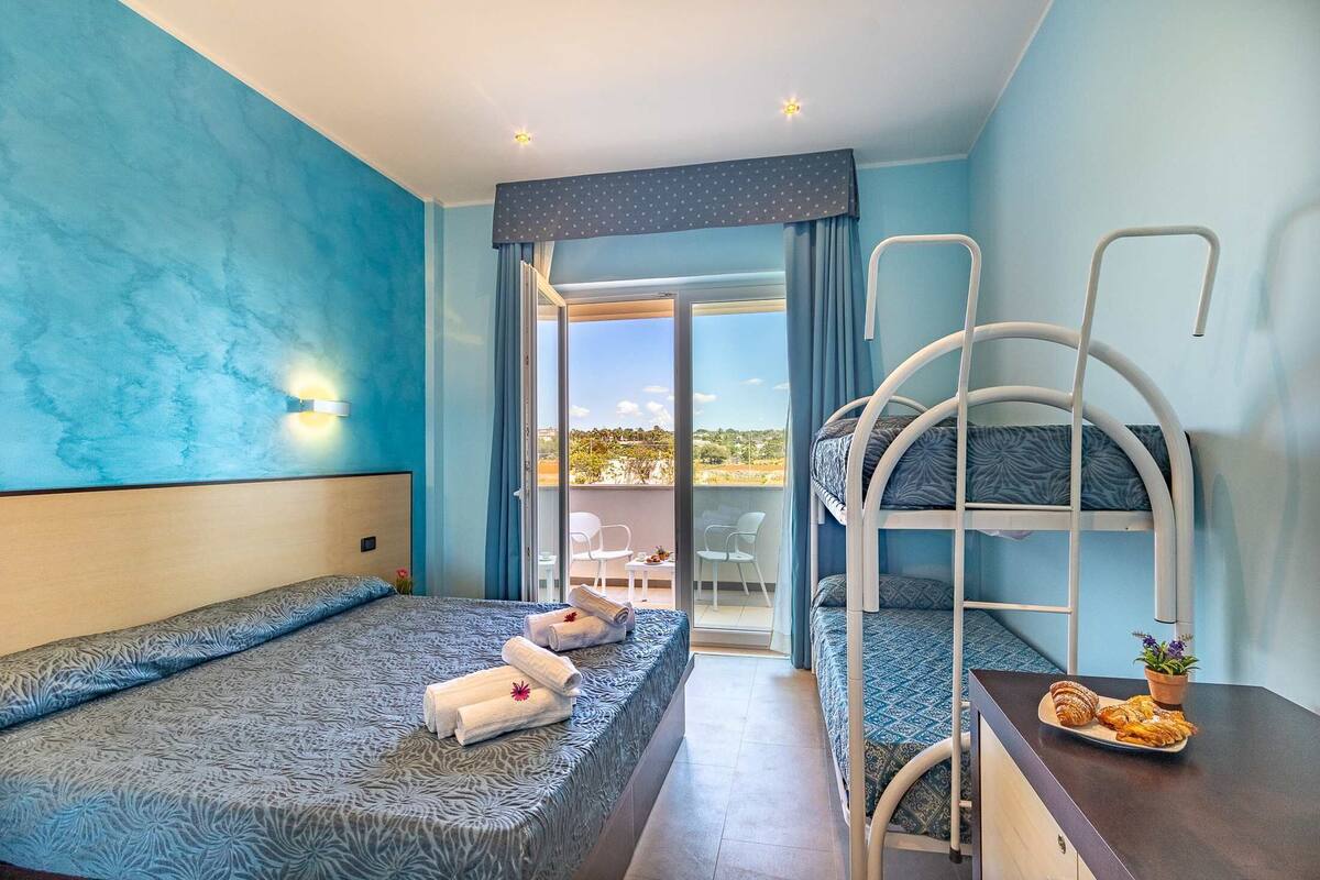A bedroom with a bunk bed, a single bed, a balcony with a table and chairs, and a view of the outdoors.