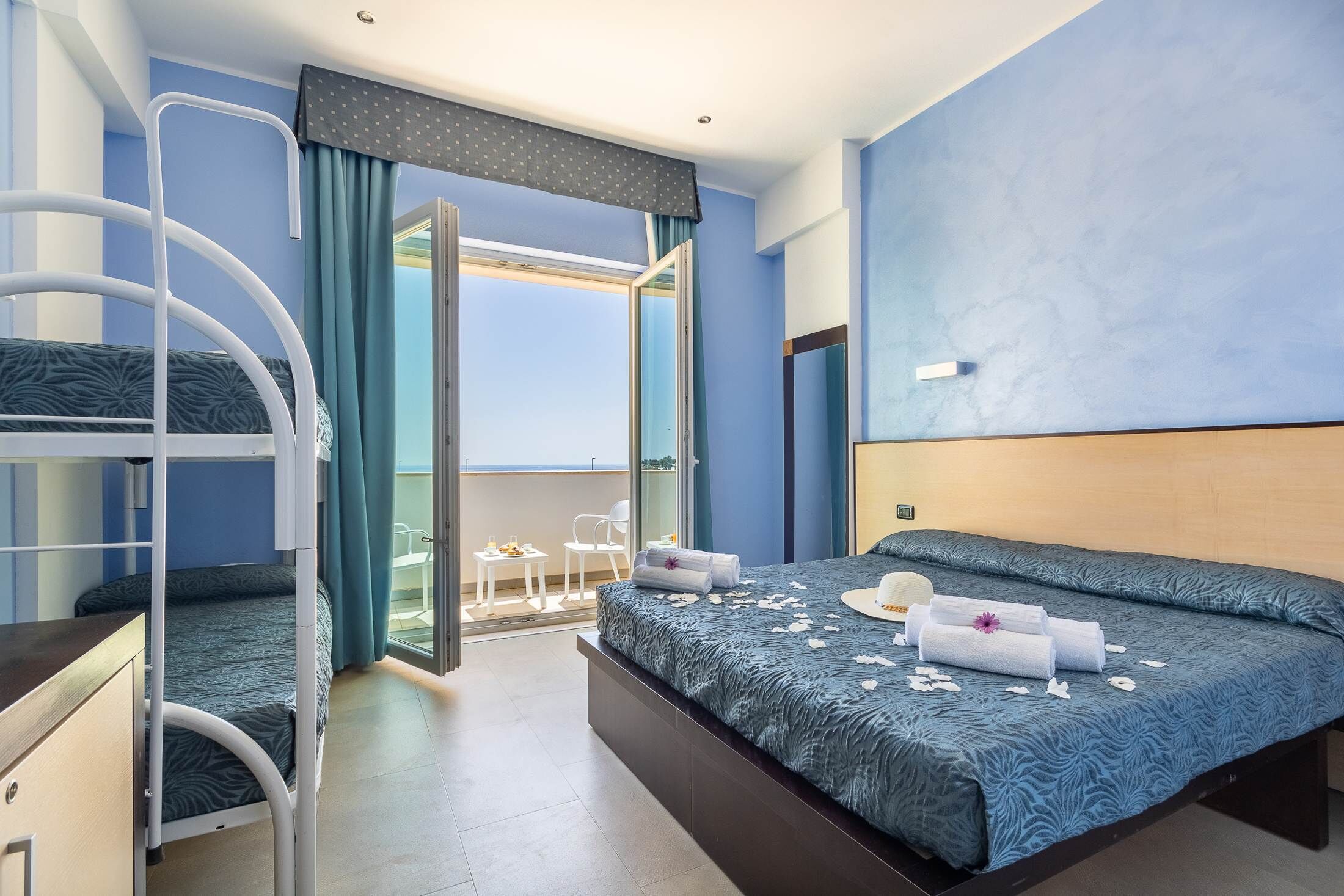 Ein modernes Hotelzimmer mit Stockbetten, einem großen Bett mit blauen, gemusterten Laken und Blick ins Freie, sowie einem Balkon mit Sitzgelegenheiten.
