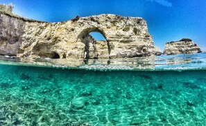 Una vista clara bajo el agua de una formación costera rocosa con un arco natural, rodeada de aguas turquesas.