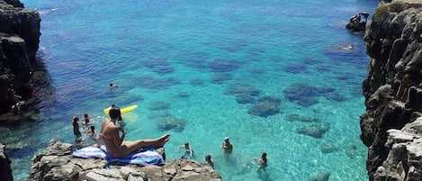 A rocky coastal area with clear blue water, people swimming and sunbathing, and a person sitting on a rock.