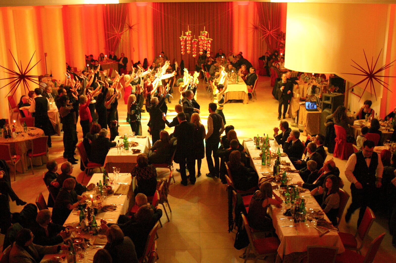 A large banquet hall with multiple round tables set for dining, a stage at the front, and a crowd of people.