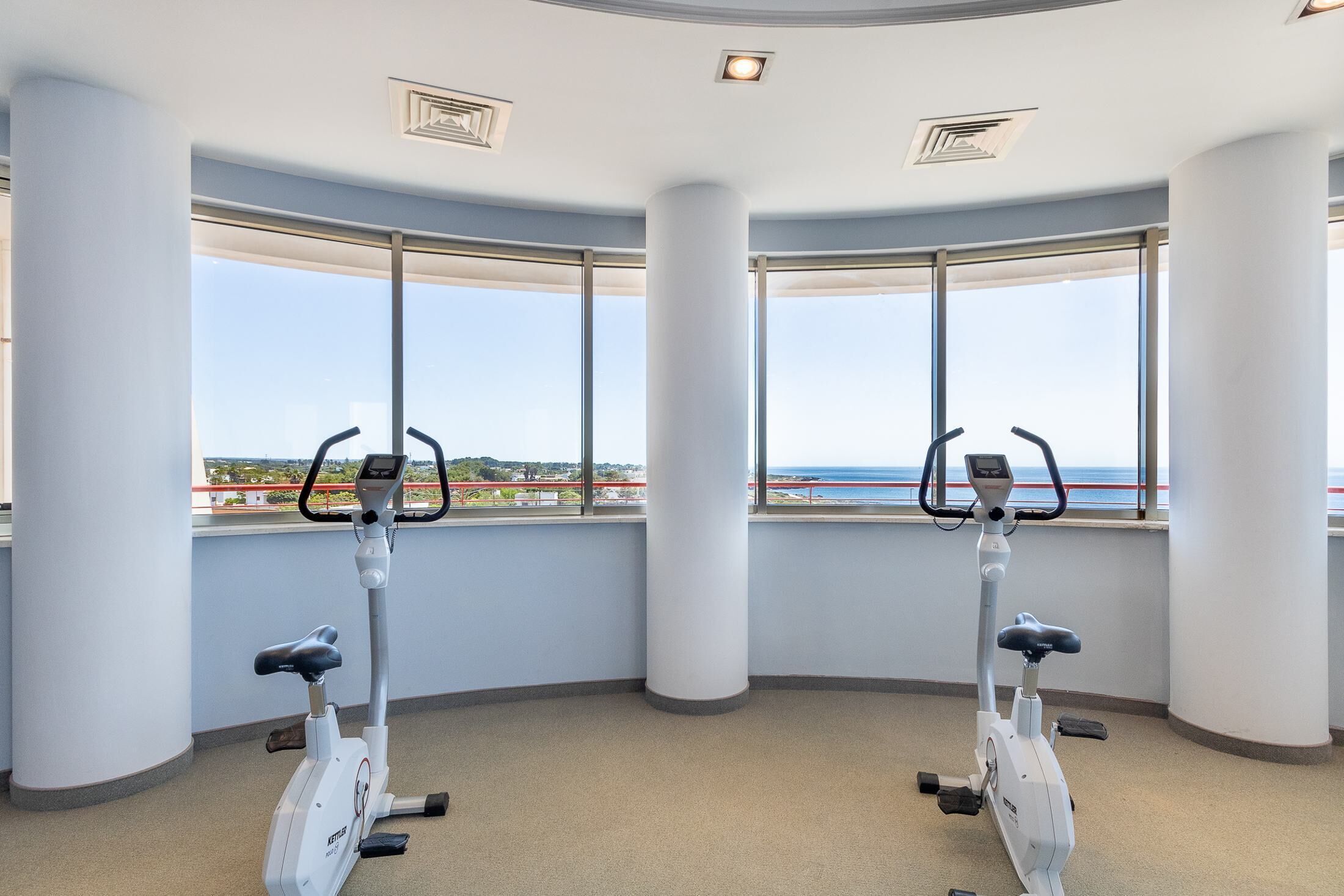 Une salle de remise en forme équipée de deux vélos d’appartement, de grandes fenêtres offrant une vue sur la mer et d’un plafond incurvé doté d’un éclairage encastré.
