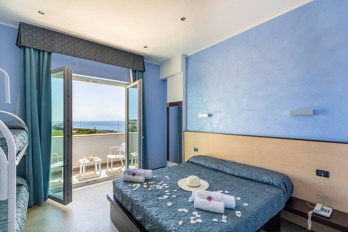 A hotel room with a bed, a balcony with a view, a phone, and a small table with a chair.