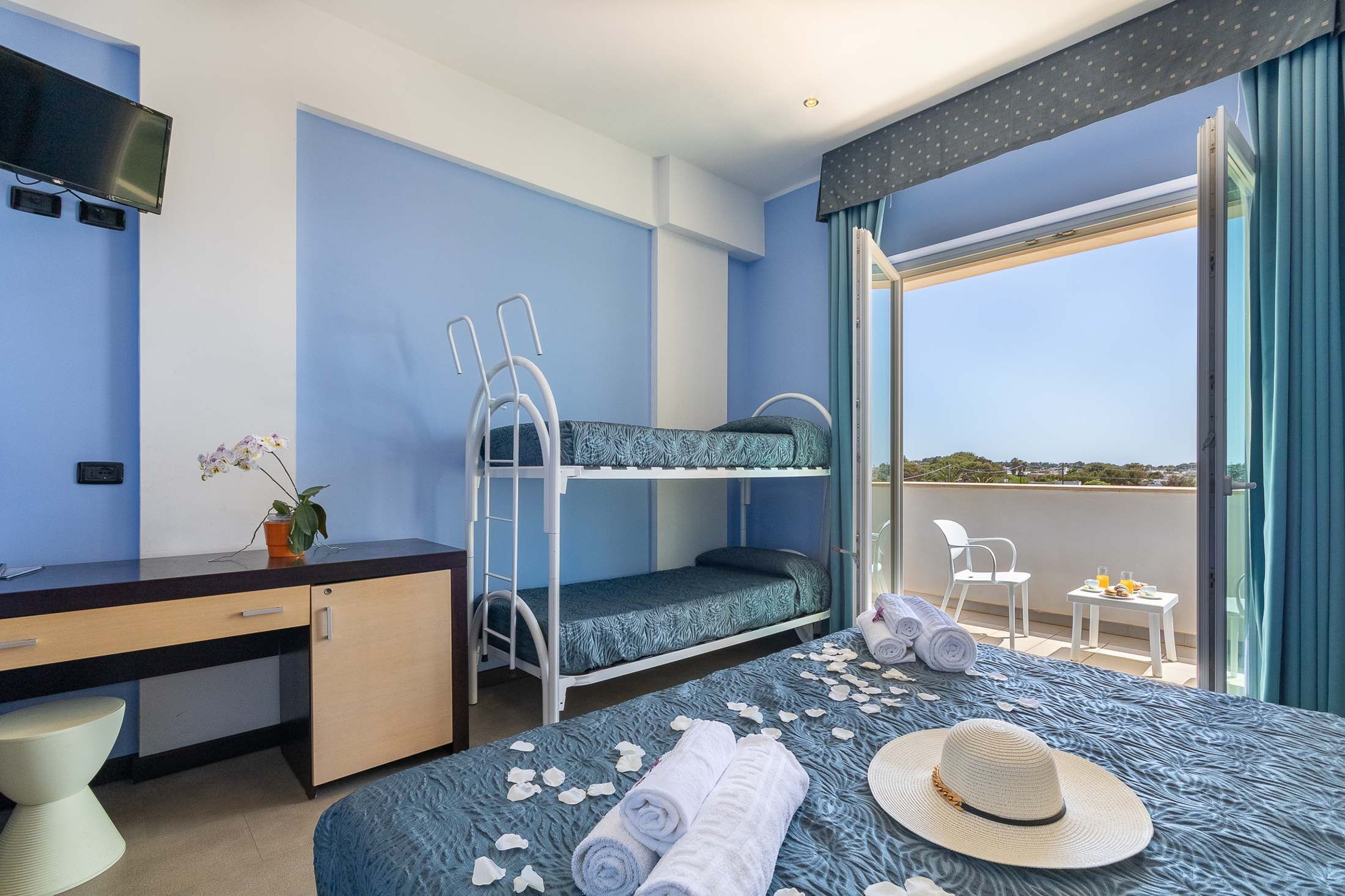 Une chambre d’hôtel avec des lits superposés, un bureau, une télévision, un balcon avec une table et des chaises, et une vue sur le paysage.